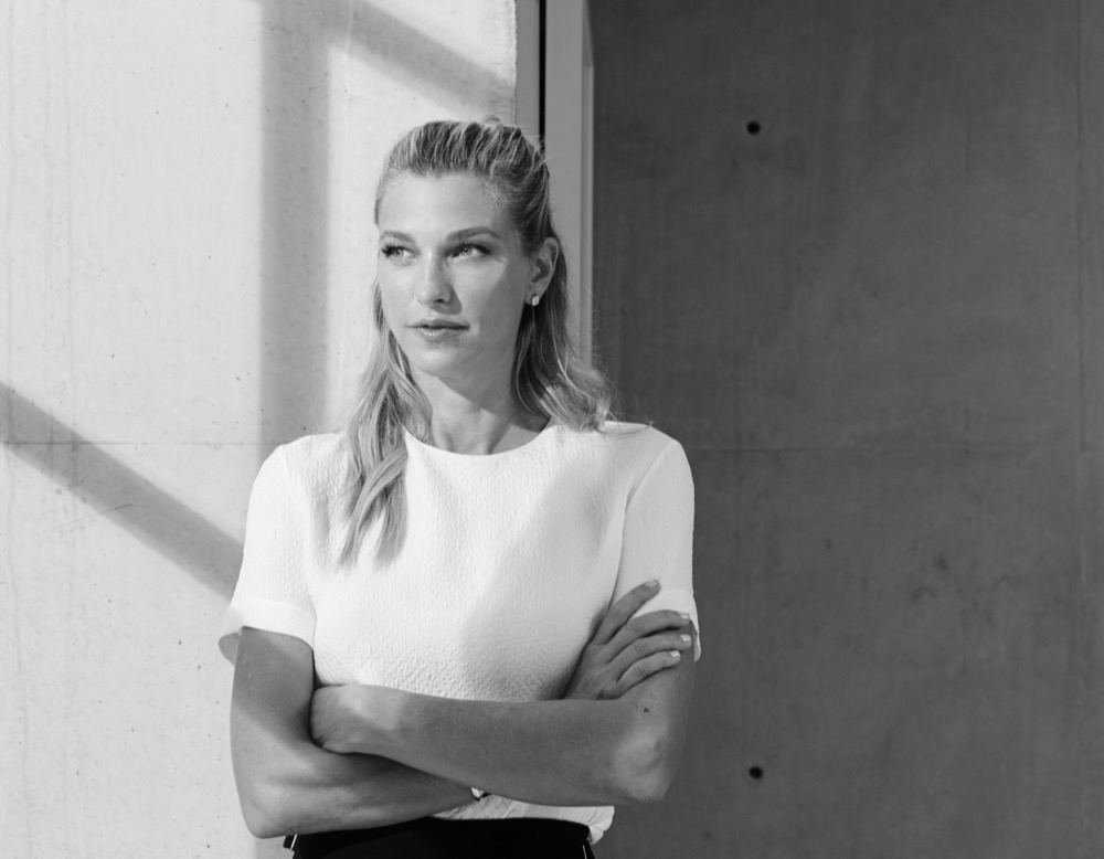 Nicole Junkermann in an ivory blazer, arms crossed, against a sunlit concrete wall