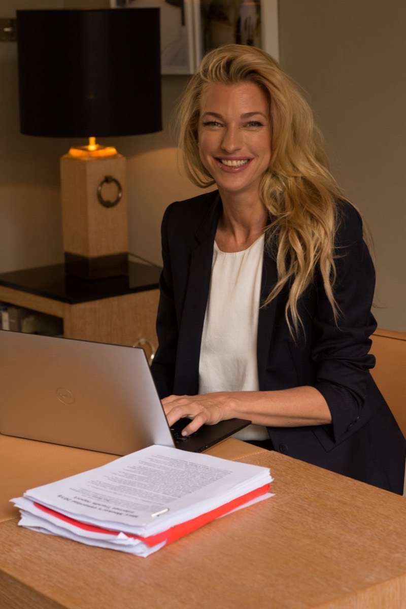 Nicole Junkermann in a navy blazer, smiling warmly at her laptop at a lamp-lit desk with documents